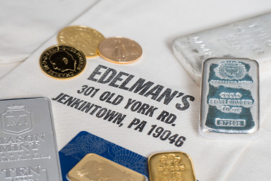 Gold and silver bullion bars and gold coins displayed on a cloth with Edelman’s business name and address